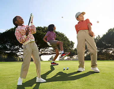 Photograph of three children playing golf on a green field with trees in the background and a clear sky.