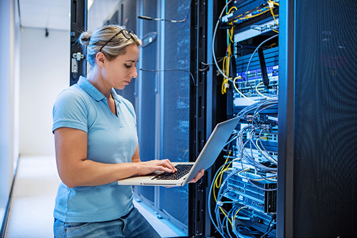 Person using a laptop while working beside network servers and cables in a data center.