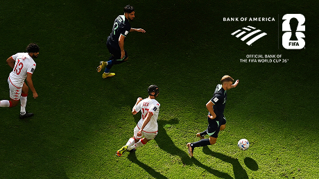 A soccer match showing four players on a green field, with two players in white jerseys and two in dark jerseys chasing a ball.