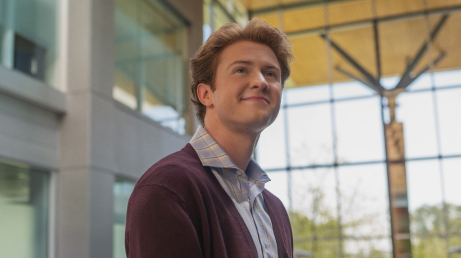Person with short hair wearing a light shirt and dark cardigan standing indoors in front of a large grid-patterned window with greenery visible outside.