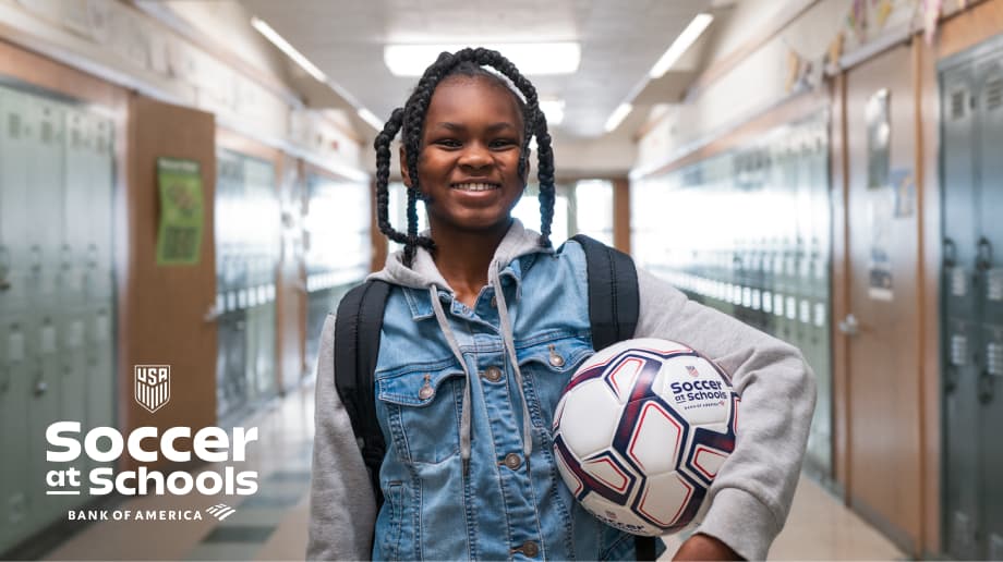 A smiling student holding a soccer ball in a school hallway 