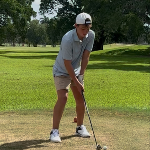 Youth golfer, Benjamin H, holds his club with his feet shoulder-width apart and his eyes directed down at the ball he’s about to hit.