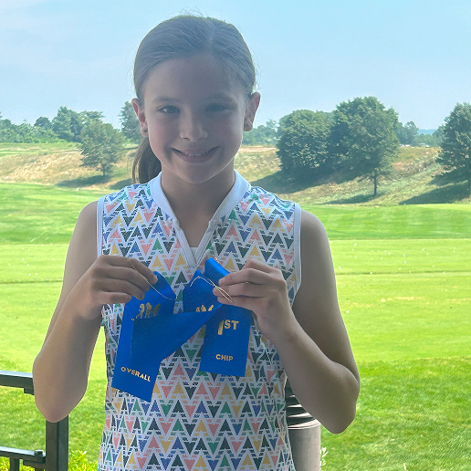 Youth golfer, Guilianna T, smiles and stands in front of a golf course while holding up awards she’s won.