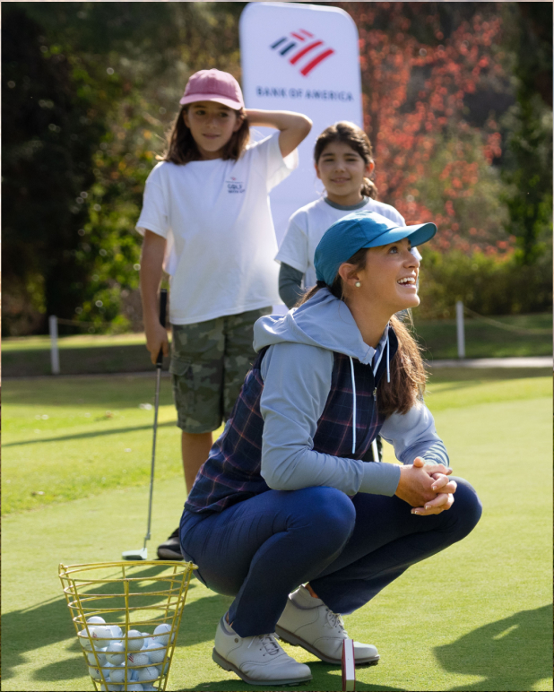 A female golf instructor, Rachel Kuehn, teaching on a golf course. She is crouched down, looking up and smiling at a student she is teaching. Children with golf clubs are standing behind her.