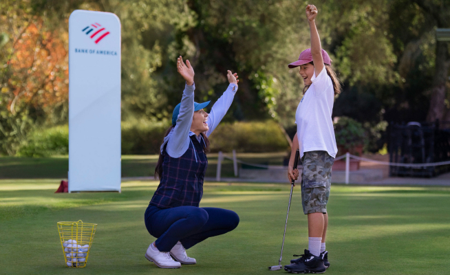 A female golf instructor, Rachel Kuehn, celebrates with one of her young students on a golf course. Both have their hands raised in the air. A Bank of America sign is in the background.