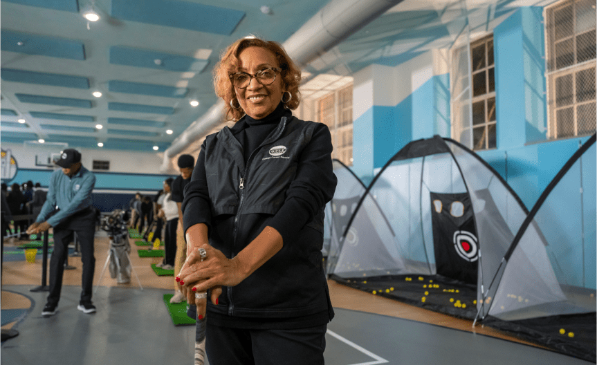 The founder of the Midnight Golf Program, Renee Fluker, stands a gymnasium where her golf clinic is being held. Teenage students in the background are practicing their swings.