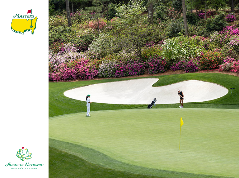 Two golfers on a putting green near a sand bunker and blooming shrubs