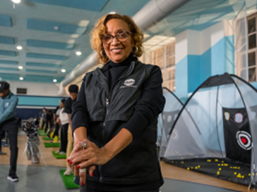 The founder of the Midnight Golf Program, Renee Fluker, stands a gymnasium where her golf clinic is being held. Teenage students in the background are practicing their swings.