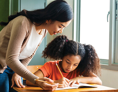 Adult helping a child with a writing activity at a classroom desk.